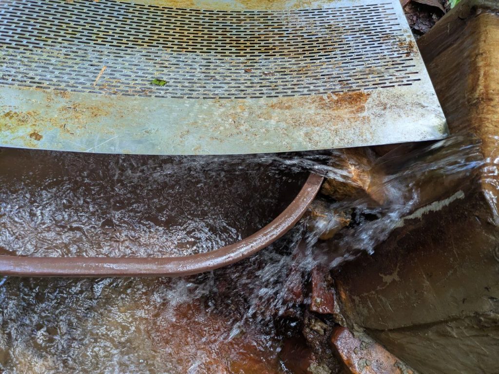 Photo of a brown bucket adjacent to a weir. There's a 20 cm gap between the weir and the bucket, such that most of the water is falling onto the ground next to the bucket (instead of inside it)
